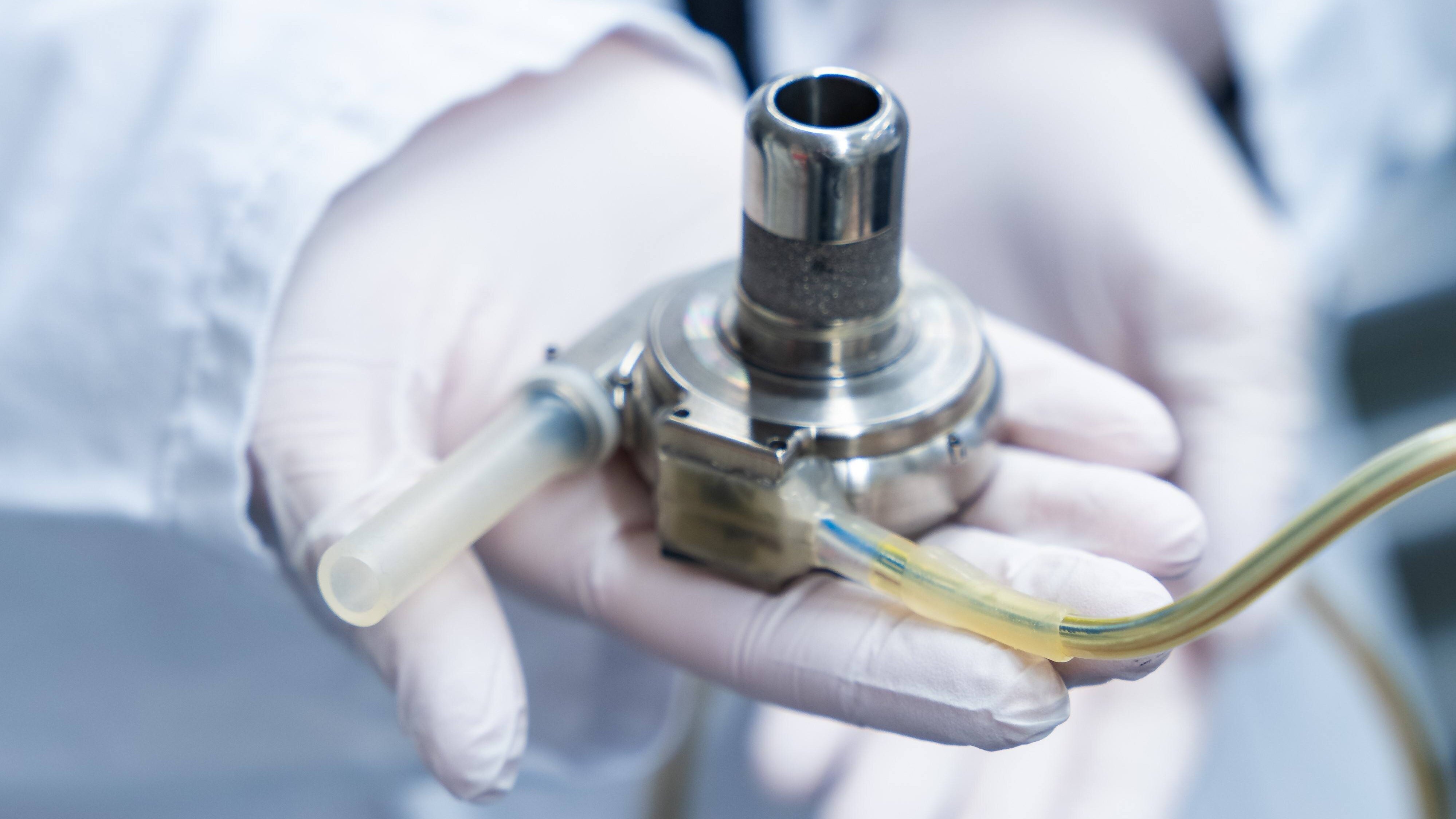 Eine Person in einem weißen Laborkittel hält ein medizinisches Gerät in der Hand. Das Gerät hat mehrere Rohre, die daran angeschlossen sind. Es ist aus Metall und scheint für medizinische Zwecke verwendet zu werden. Die Person trägt auch Einweghandschuhe.
