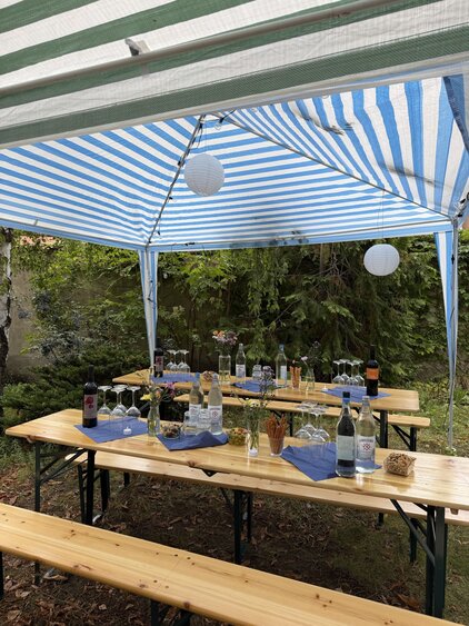 Ein festlich gedeckter Tisch unter einem blauen und weißen Zelt. Es gibt Gläser, Wasserflaschen und Weinflaschen, sowie kleine Snacks. Der Hintergrund ist von grünen Büschen umgeben, die eine einladende Atmosphäre schaffen.