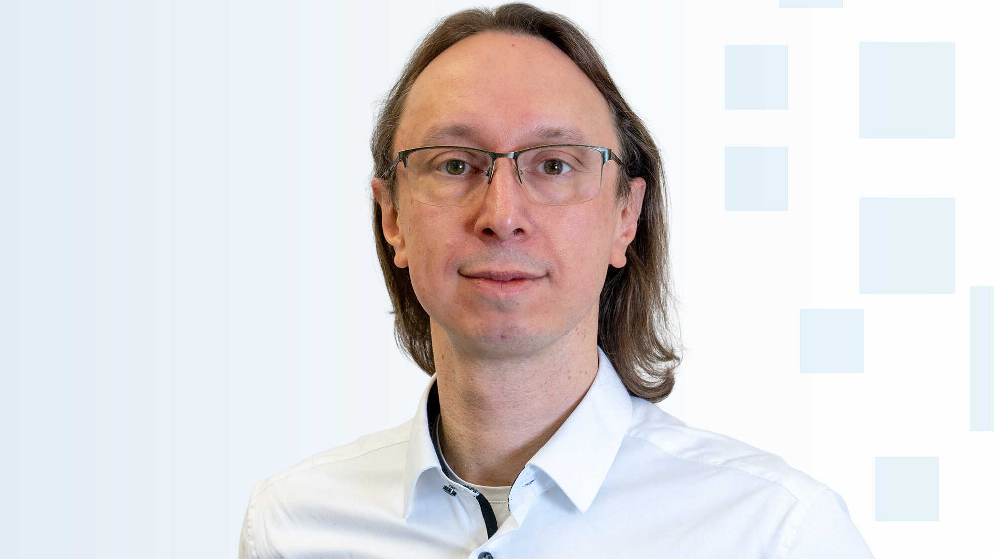 Ein Mann mit langen, braunen Haaren und einer Brille trägt ein weißes Hemd. Er steht vor einem hellen, einfarbigen Hintergrund mit grafischen Mustern. Sein Gesichtsausdruck ist freundlich und neutral.