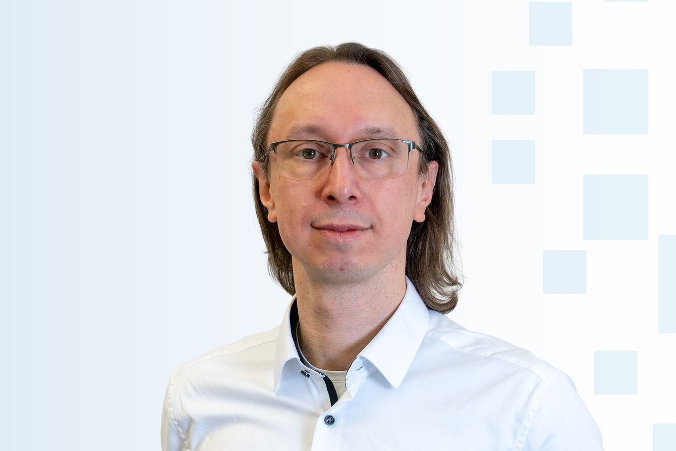 Ein Mann mit langen, braunen Haaren und einer Brille trägt ein weißes Hemd. Er steht vor einem hellen, einfarbigen Hintergrund mit grafischen Mustern. Sein Gesichtsausdruck ist freundlich und neutral.