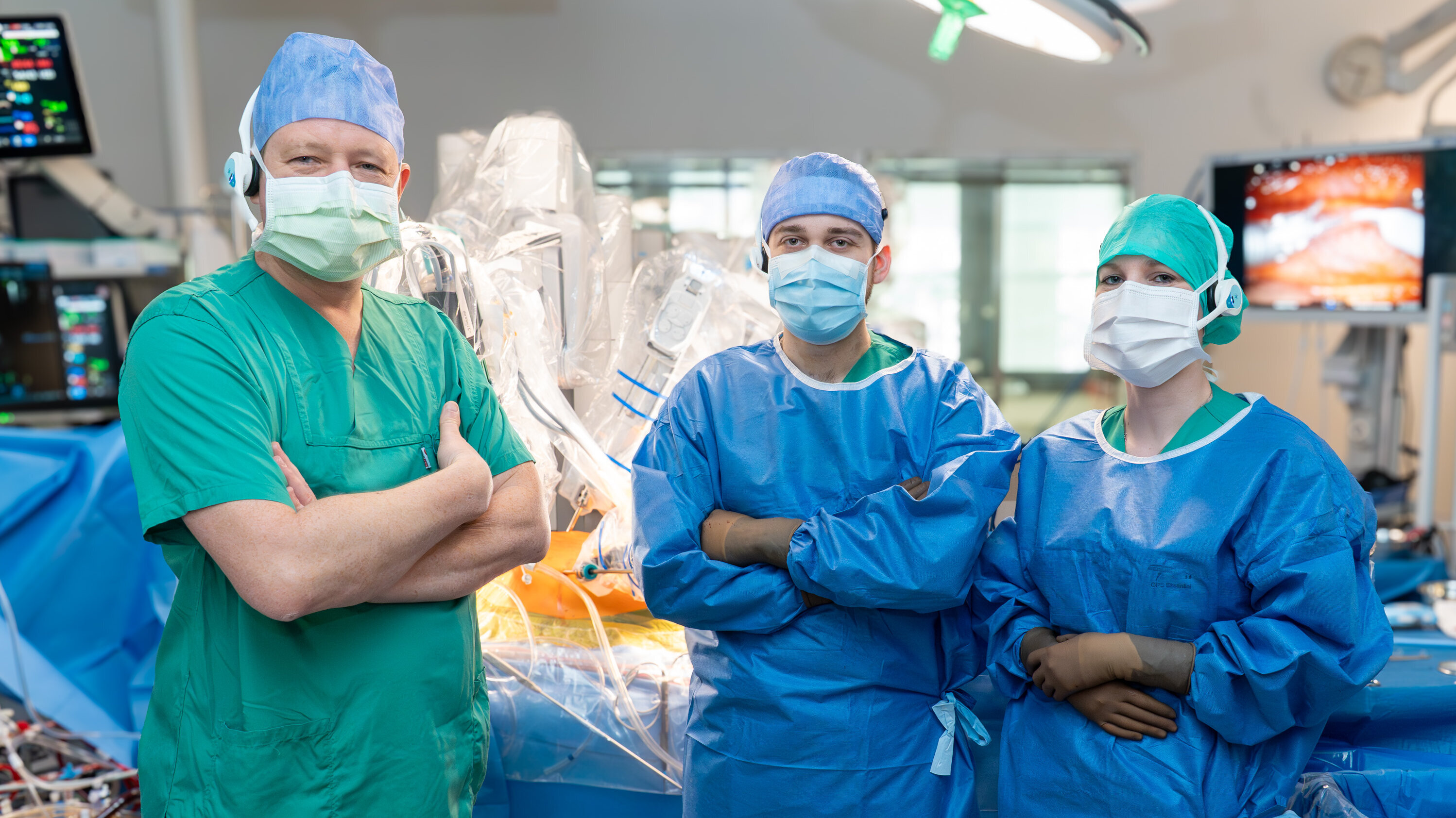 Drei medizinische Fachkräfte stehen in einem Operationssaal. Sie tragen medizinische Kleidung, Masken und Kappen. Im Hintergrund sind chirurgische Geräte und Instrumente sichtbar. Die Atmosphäre vermittelt Professionalität und Vorbereitung für einen Eingriff.