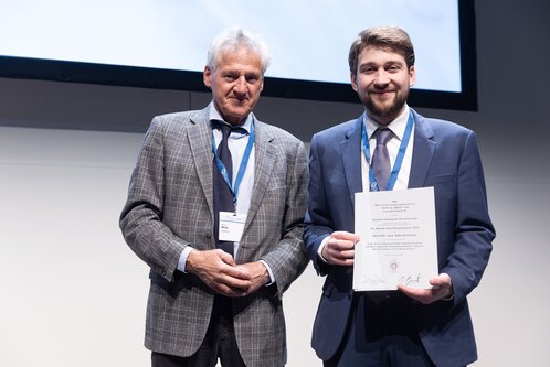 Zwei Männer stehen nebeneinander auf einer Bühne. Der ältere Mann, in einem karierten Anzug, lächelt und hält eine Urkunde. Der jüngere Mann, gekleidet in einen Anzug mit einer Krawatte, zeigt stolz die Urkunde, die er gerade erhalten hat.
