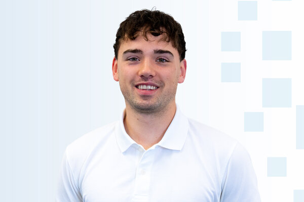 Ein junger Mann mit kurzen, lockigen Haaren trägt ein weißes Polohemd. Er lächelt und steht vor einem hellen, einfarbigen Hintergrund mit dezenten geometrischen Mustern.