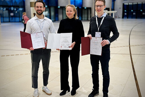 Drei Personen stehen in einem großen, hellen Raum und halten Zertifikate in den Händen. Die beiden Männer tragen unterschiedliche Outfits, einer in heller Kleidung und der andere in dunkler. Die Frau in der Mitte trägt schwarze Kleidung und lächelt. Alle drei scheinen stolz auf ihre Auszeichnungen zu sein.
