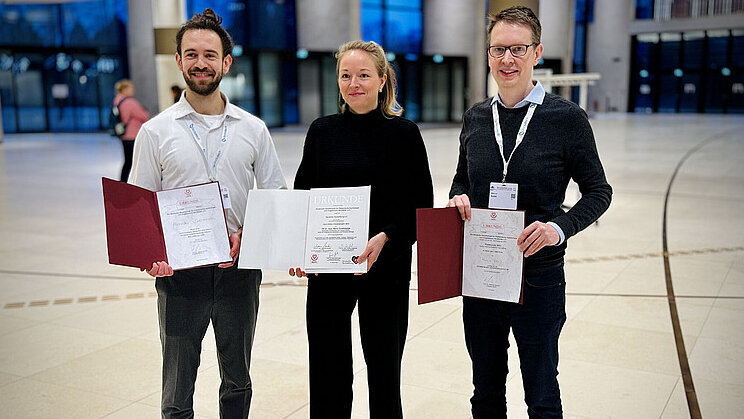 Drei Personen stehen in einem großen, hellen Raum und halten Zertifikate in den Händen. Die beiden Männer tragen unterschiedliche Outfits, einer in heller Kleidung und der andere in dunkler. Die Frau in der Mitte trägt schwarze Kleidung und lächelt. Alle drei scheinen stolz auf ihre Auszeichnungen zu sein.