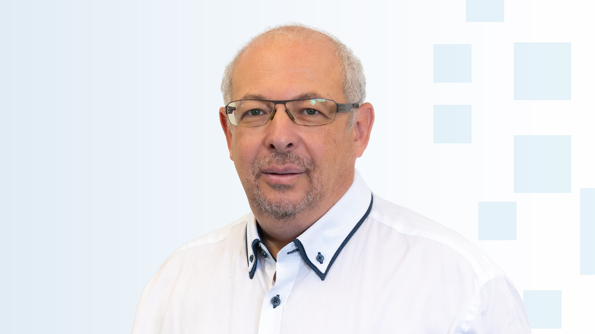 Ein Mann mit Brille und einem weißen Hemd lächelt in die Kamera. Der Hintergrund ist hell und einfach gestaltet.