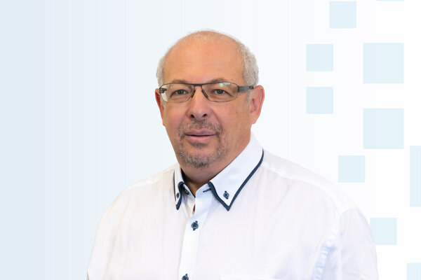 Ein Mann mit Brille und einem weißen Hemd lächelt in die Kamera. Der Hintergrund ist hell und einfach gestaltet.