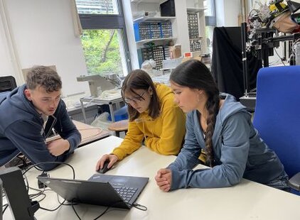 Drei junge Menschen sitzen an einem Tisch und schauen gemeinsam auf einen Laptop. Sie scheinen sich über den Inhalt des Bildschirms auszutauschen. Die Atmosphäre wirkt konzentriert und kooperativ.