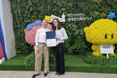 Ein Mann und eine Frau stehen inmitten einer grünen Blumenwand. Die Frau hält ein Zertifikat in der Hand. Beide lächeln freundlich. Hinter ihnen sind farbenfrohe, blumenverzierte Figuren zu sehen.