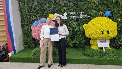Ein Mann und eine Frau stehen inmitten einer grünen Blumenwand. Die Frau hält ein Zertifikat in der Hand. Beide lächeln freundlich. Hinter ihnen sind farbenfrohe, blumenverzierte Figuren zu sehen.