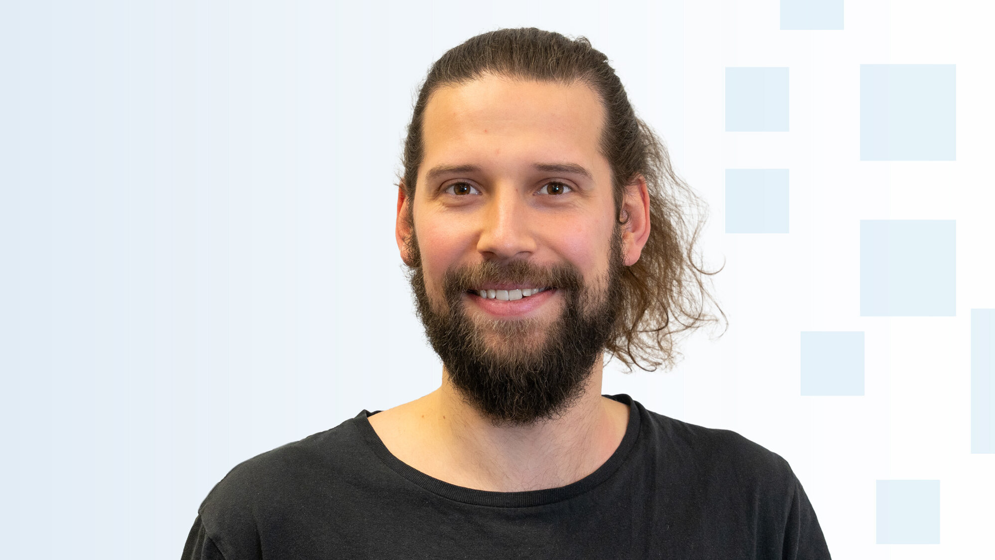 Ein junger Mann mit einem langen, lockigen Bart und langen, ungekämmten Haaren lächelt in die Kamera. Er trägt ein schwarzes T-Shirt und hat einen freundlichen Ausdruck. Der Hintergrund ist hellblau mit einfachen grafischen Elementen.