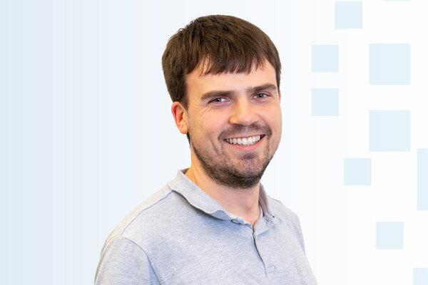 Ein junger Mann mit kurzen, braunen Haaren trägt ein graues Polohemd und lächelt. Der Hintergrund ist hell und enthält grafische Elemente in Form von hellen rechteckigen Formen.