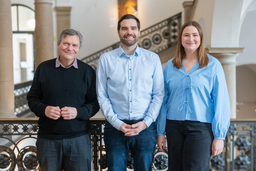 Drei Personen stehen zusammen vor einem geländerten Geländer in einem Innenraum. Der Mann links trägt einen schwarzen Pullover, der Mann in der Mitte ein hellblaues Hemd, und die Frau rechts trägt eine hellblaue Bluse. Sie lächeln freundlich in die Kamera.