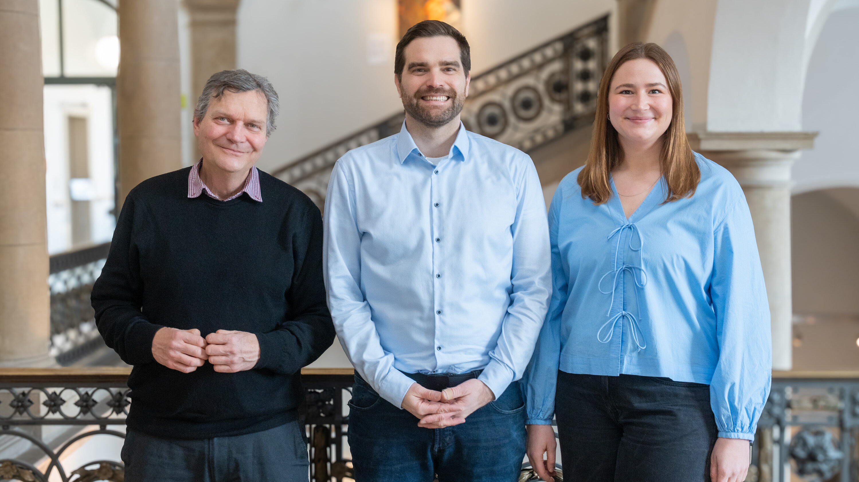 Drei Personen stehen zusammen vor einem geländerten Geländer in einem Innenraum. Der Mann links trägt einen schwarzen Pullover, der Mann in der Mitte ein hellblaues Hemd, und die Frau rechts trägt eine hellblaue Bluse. Sie lächeln freundlich in die Kamera.