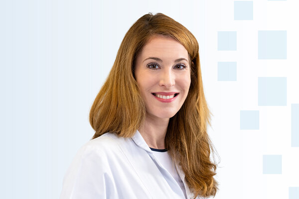 Porträt einer lächelnden Frau mit langen, rotbraunen Haaren, die einen weißen Laborkittel trägt. Der Hintergrund ist sanft hellblau mit geometrischen Formen. Sie strahlt Freundlichkeit und Professionalität aus.