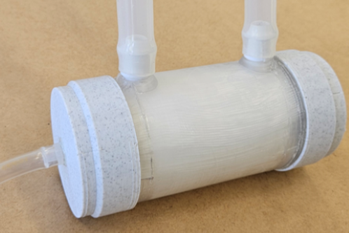 Ein zylindrisches Gerät mit zwei Schläuchen, das aus einem hellen Material gefertigt ist. Es hat an beiden Enden runde Kappen und ist auf einer hellbraunen Oberfläche platziert. Die Schläuche sind transparent und verlaufen von der Oberseite des Geräts.