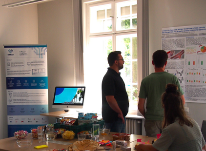 Zwei Personen stehen im Vordergrund und betrachten ein Plakat an der Wand. Auf dem Tisch im Hintergrund sind Snacks und Getränke platziert. Ein Computerbildschirm zeigt ein Bild an. Im Raum gibt es Fenster mit Tageslicht.