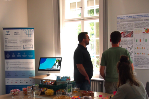 Zwei Personen stehen im Vordergrund und betrachten ein Plakat an der Wand. Auf dem Tisch im Hintergrund sind Snacks und Getränke platziert. Ein Computerbildschirm zeigt ein Bild an. Im Raum gibt es Fenster mit Tageslicht.