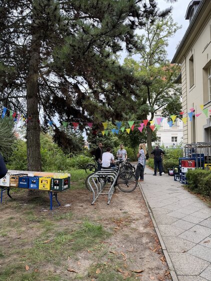 Eine Gruppe von Menschen steht in einem Garten, umgeben von Bäumen und bunten Wimpeln. Einige Personen sitzen auf Fahrrädern, während andere sich unterhalten. Im Hintergrund sind bunte Kisten zu sehen, die auf einem Tisch gestapelt sind. Die Atmosphäre wirkt fröhlich und gesellig.